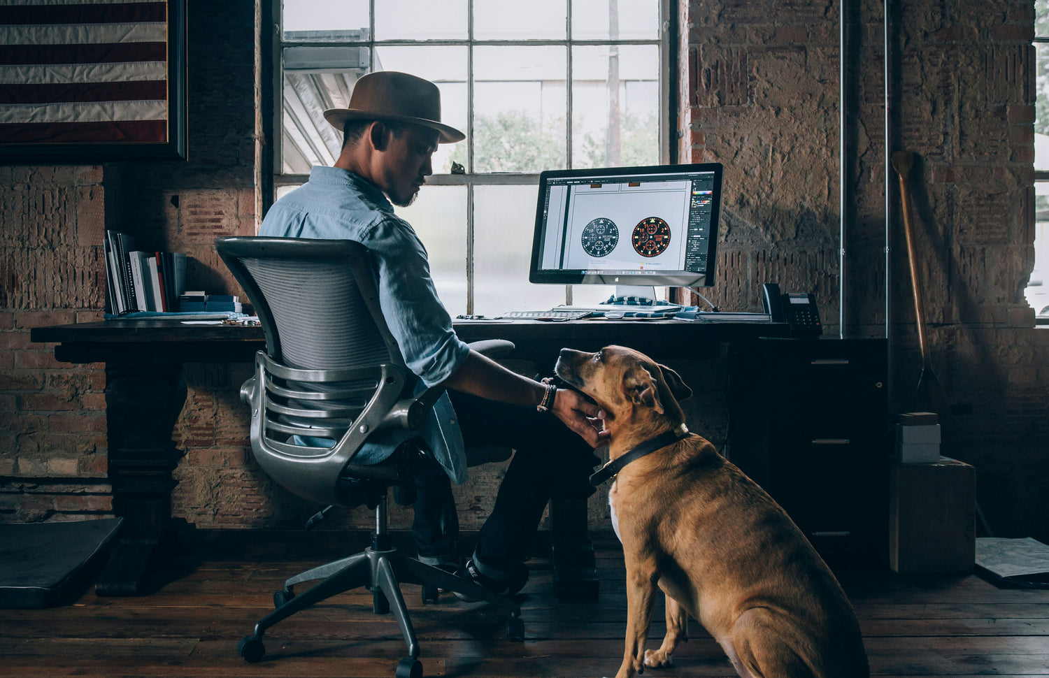 a man sitting in an office in front of a desk while scratching the chin of a dog next to him